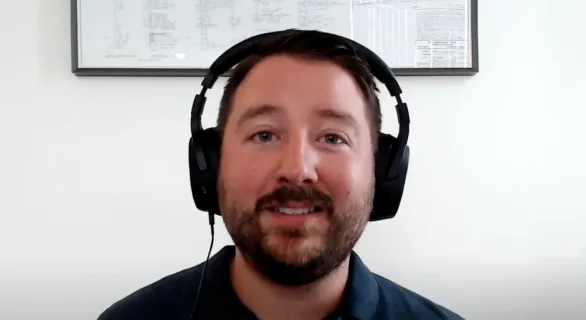 Man with beard wearing black headphones smiling in front of a white wall with framed documents.
